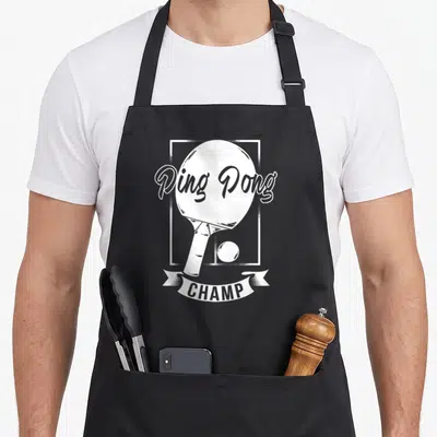 Pong champ table apron - White ping pong paddle and ball in a frame with CHAMP banner text — shown on a black cotton canvas apron against a white background