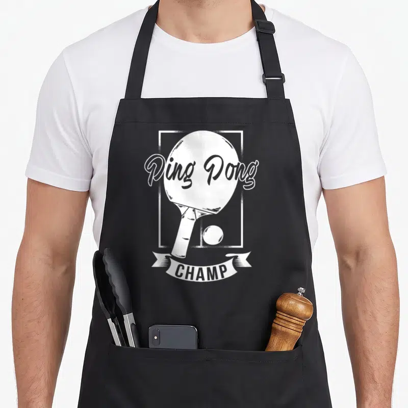 Pong champ table apron - White ping pong paddle and ball in a frame with CHAMP banner text — shown on a black cotton canvas apron against a white background
