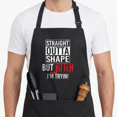Outta shapes apron - White rectangle frame with STRAIGHT OUTTA SHAPE text and red BITCH script below — shown on a black cotton canvas apron against a white background