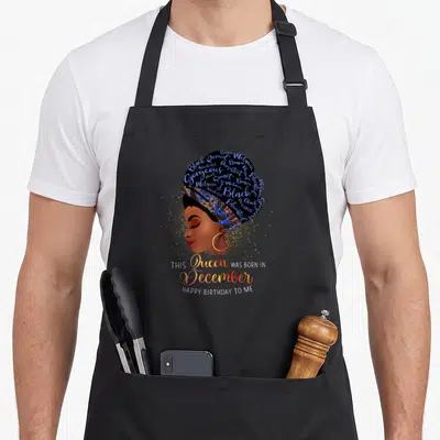 December afro birthday apron - Profile of woman with blue word art headwrap and gold glitter celebrating December — shown on a black cotton canvas apron against a white background