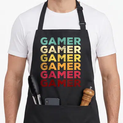 70s video game apron - Six stacked GAMER words in teal, yellow, orange, pink, and red with cracked texture — shown on a black cotton canvas apron against a white background