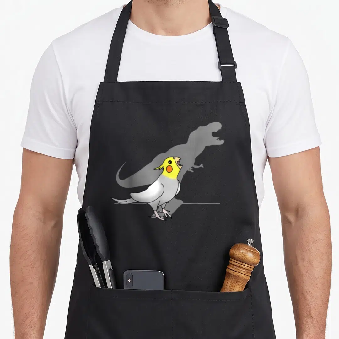 Screaming t-rex apron - Cartoon cockatiel with yellow head and grey wings in front of a faint dinosaur silhouette — shown on a black cotton canvas apron against a white background