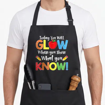 Test show teacher apron - Stacked text with red apple replacing O in GLOW, two pencils flanking What you, in white red — shown on a black cotton canvas apron against a white background