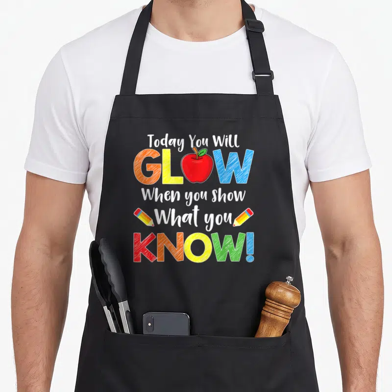 Test show teacher apron - Stacked text with red apple replacing O in GLOW, two pencils flanking What you, in white red — shown on a black cotton canvas apron against a white background