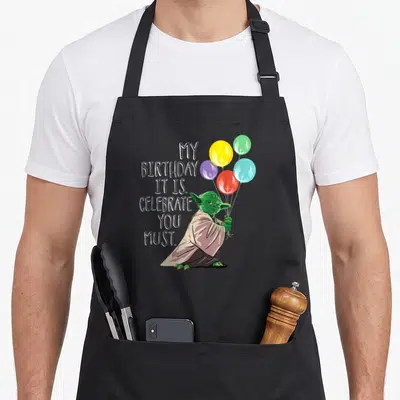 Yoda celebrate birthday apron - Cartoon Yoda holding five colorful balloons next to stacked white text reading MY — shown on a black cotton canvas apron against a white background