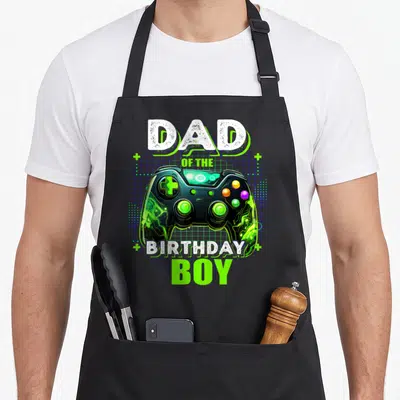 Birthday video game apron for women - Game controller on blue grid with DAD OF THE BIRTHDAY BOY text and four plus signs — shown on a black cotton canvas apron against a white background