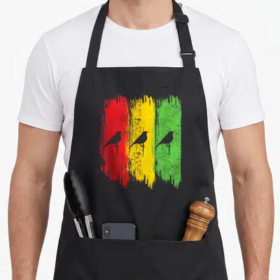 Jamaica reggae apron - Three grey birds overlaid on red, yellow, and green vertical brushstrokes — shown on a black cotton canvas apron against a white background