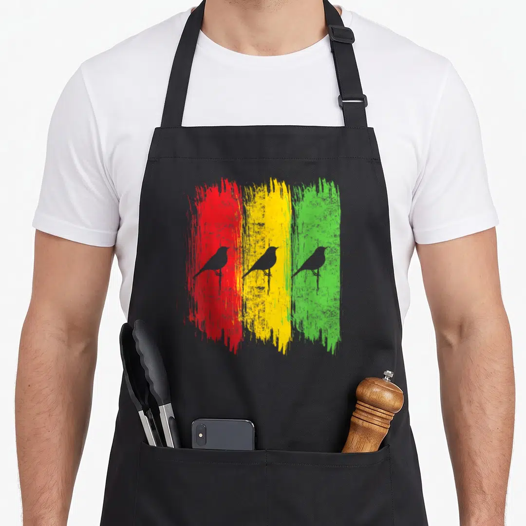 Jamaica reggae apron - Three grey birds overlaid on red, yellow, and green vertical brushstrokes — shown on a black cotton canvas apron against a white background