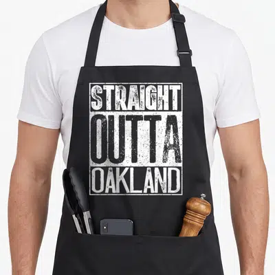 Straight apron or apron - Distressed white text reading STRAIGHT OUTTA OAKLAND stacked vertically in a rectangular — shown on a black cotton canvas apron against a white background