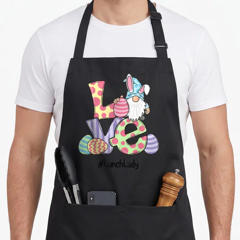 Easter lunch lady apron - Cartoon gnome with bunny ears holding egg behind colorful LOVE letters and #lunchlandy text — shown on a black cotton canvas apron against a white background