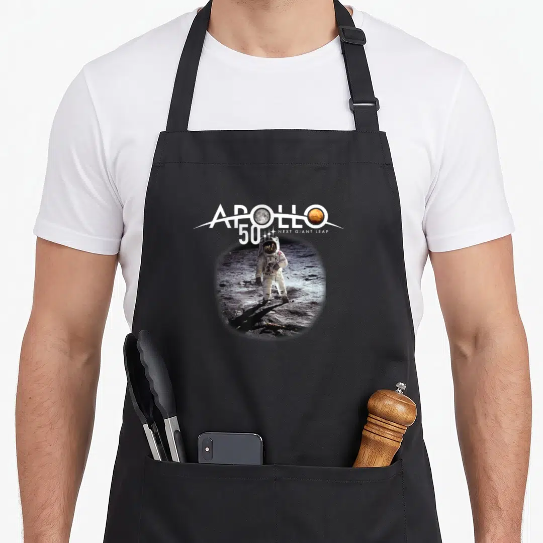 Walk apollo apron - APOLLO 50 Next Giant Leap text with astronaut photo on moon surface — shown on a black cotton canvas apron against a white background