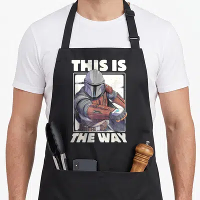 Way star apron - Cartoon Mandalorian in white frame with This Is The Way text — shown on a black cotton canvas apron against a white background