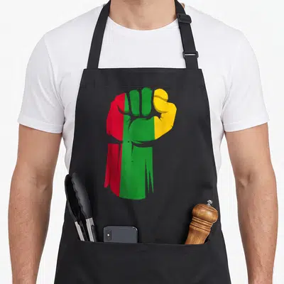 Fist african print apron - Raised fist filled with red, green, yellow vertical stripes on black background — shown on a black cotton canvas apron against a white background