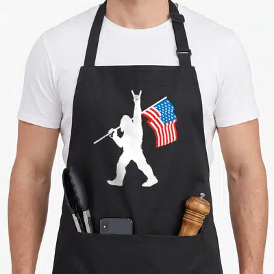 Flag bigfoot apron - White Bigfoot silhouette holding waving USA flag and making rock sign with red white blue colors — shown on a black cotton canvas apron against a white background