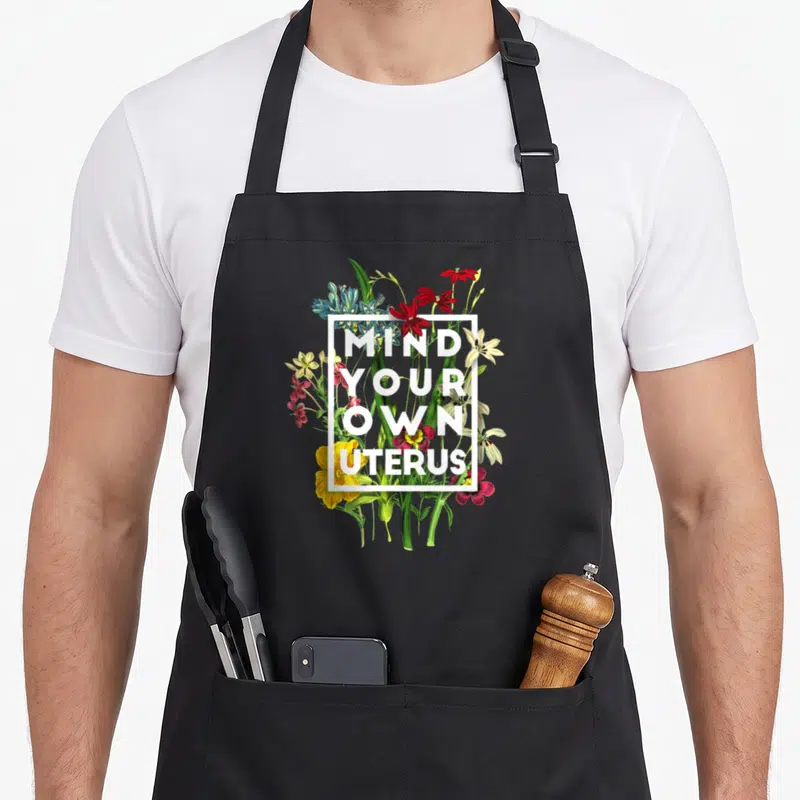 Mind floral apron - Floral cluster with white square frame containing stacked mind your own uterus text — shown on a black cotton canvas apron against a white background