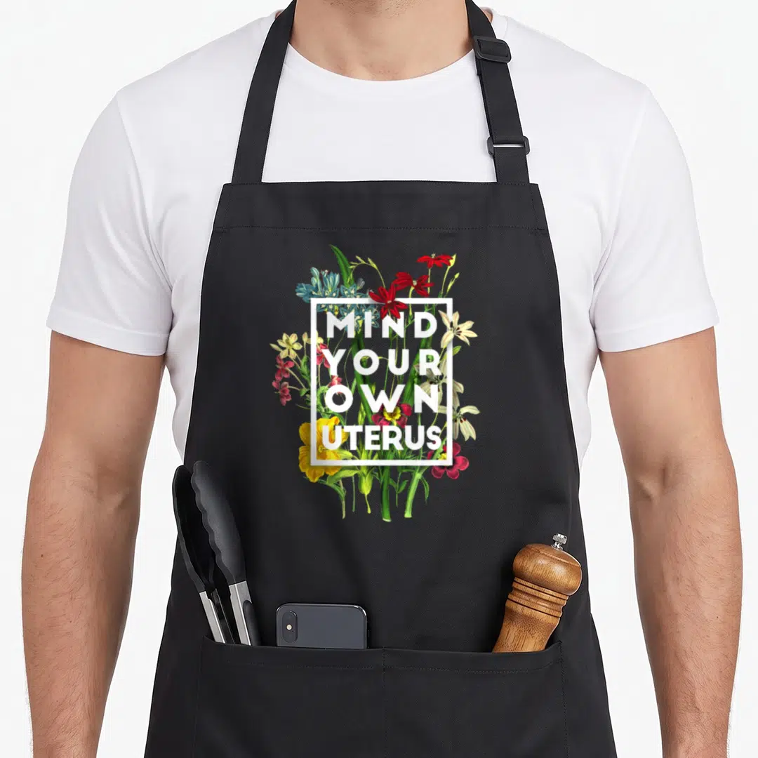 Mind floral apron - Floral cluster with white square frame containing stacked mind your own uterus text — shown on a black cotton canvas apron against a white background