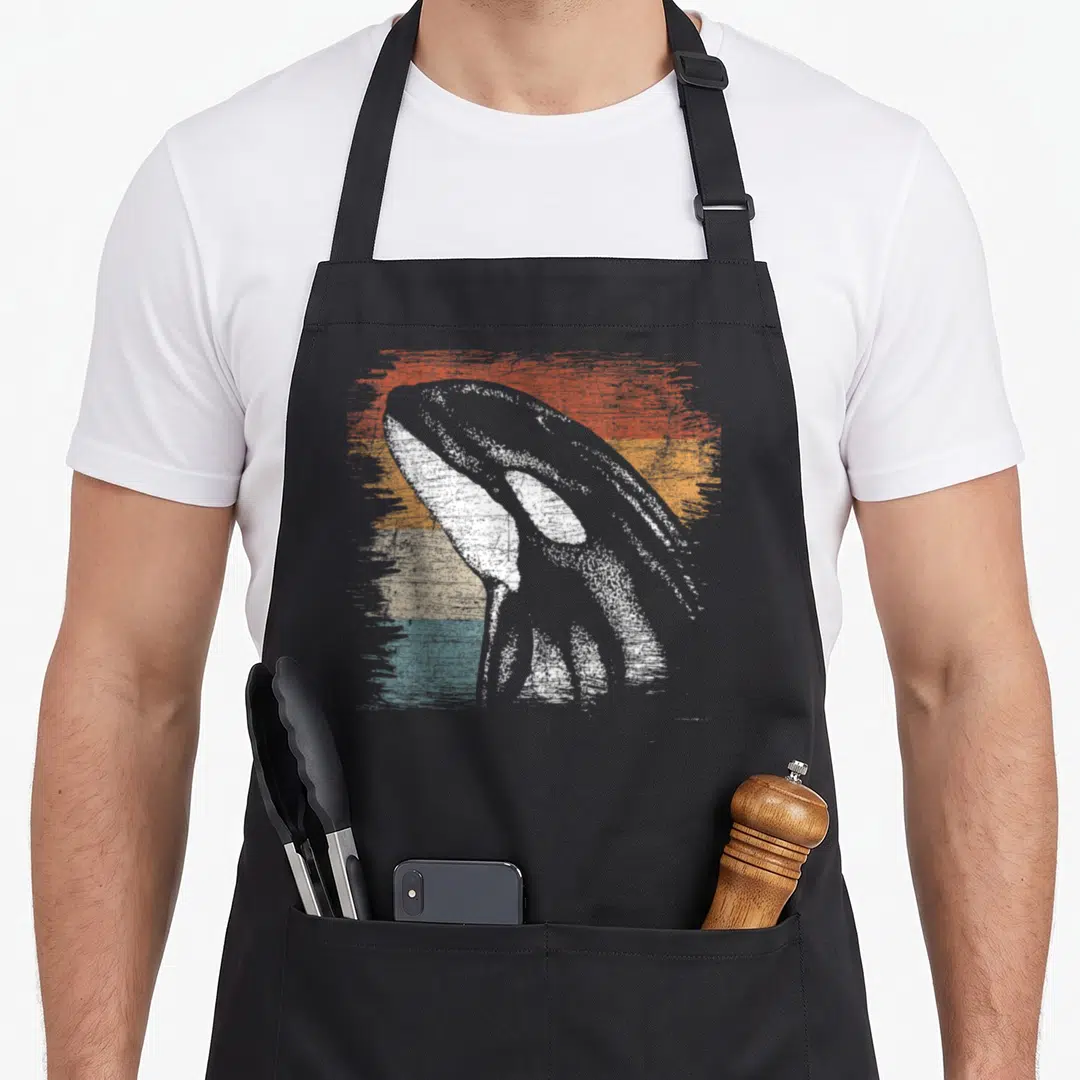 Funny killer whale apron - Distressed white whale over red orange beige and teal horizontal stripes — shown on a black cotton canvas apron against a white background
