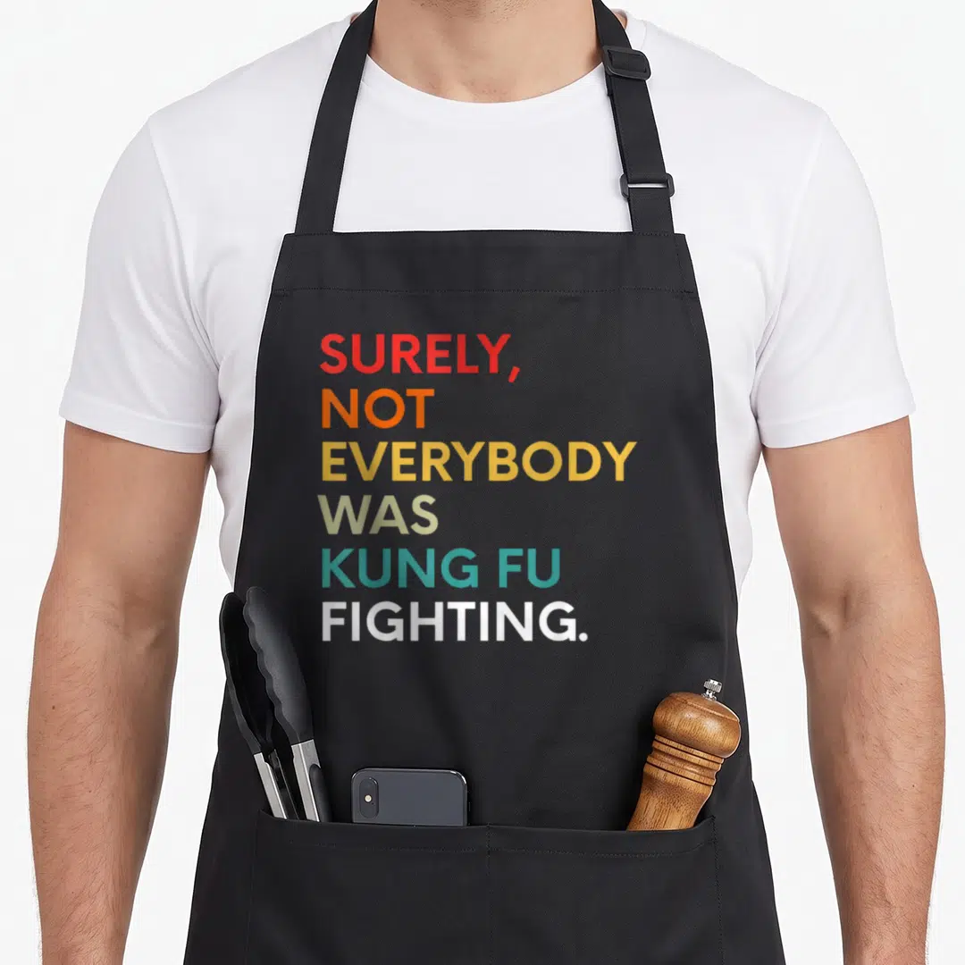 Everybody kung apron - Stacked colorful text reading SURELY NOT EVERYBODY WAS KUNG FU FIGHTING on grey background — shown on a black cotton canvas apron against a white background