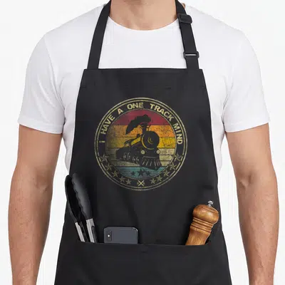 Mind track apron - Retro steam locomotive over multicolored stripes with arched text and RR symbol — shown on a black cotton canvas apron against a white background