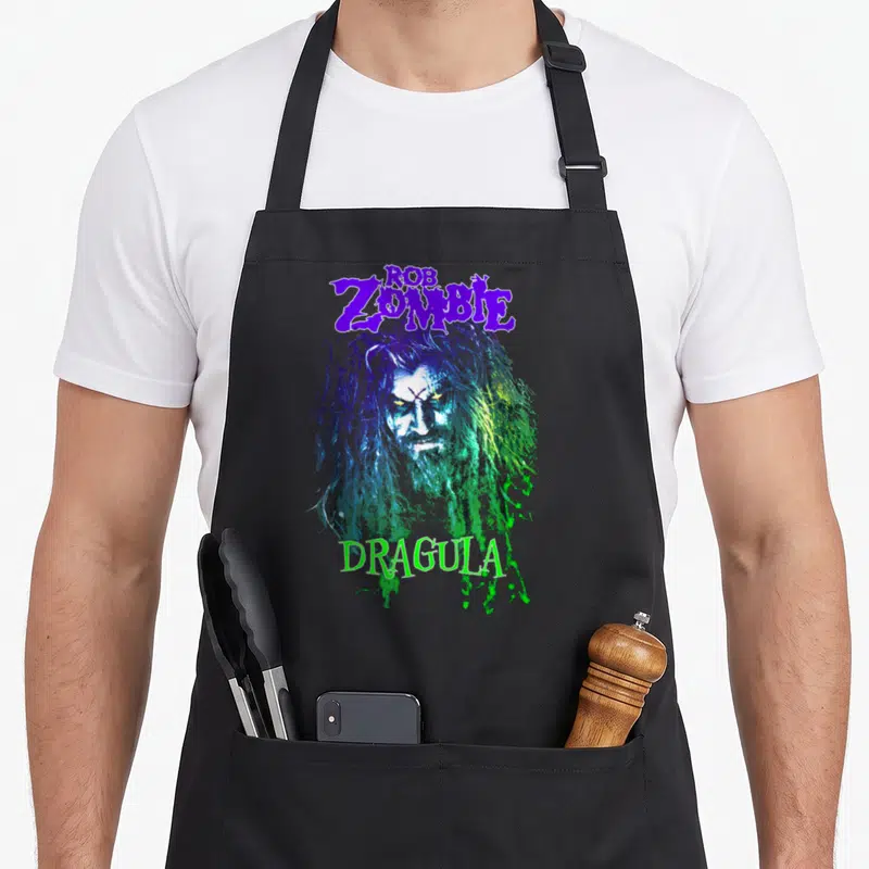 Rob zombie apron - Distressed portrait of a man with purple and green hair flanked by text reading Rob Zombie Dragula — shown on a black cotton canvas apron against a white background