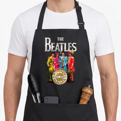 Sgt pepper apron - The Beatles standing in colorful uniforms behind a drum with SGT PEPPER'S LONELY HEARTS CLUB BAND — shown on a black cotton canvas apron against a white background