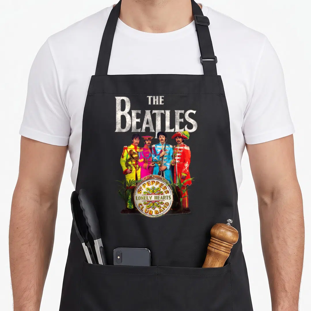 Sgt pepper apron - The Beatles standing in colorful uniforms behind a drum with SGT PEPPER'S LONELY HEARTS CLUB BAND — shown on a black cotton canvas apron against a white background