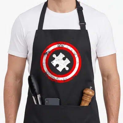 Captain superhero apron - Distressed white puzzle piece inside two concentric red and white circles — shown on a black cotton canvas apron against a white background