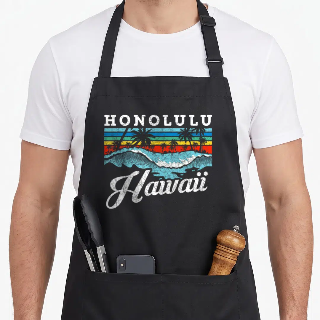 Wave hawaiian apron - HONOLULU text above striped sunset with palm trees and wave, Hawaii script below — shown on a black cotton canvas apron against a white background