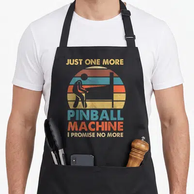 Pub pinball apron - Retro sun graphic with person playing pinball and sarcastic text about arcade addiction — shown on a black cotton canvas apron against a white background