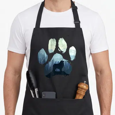 Pyrenee paw dog apron - Paw print silhouette containing a bear standing on a rock in a blue forest landscape — shown on a black cotton canvas apron against a white background