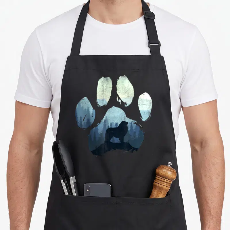 Pyrenee paw dog apron - Paw print silhouette containing a bear standing on a rock in a blue forest landscape — shown on a black cotton canvas apron against a white background