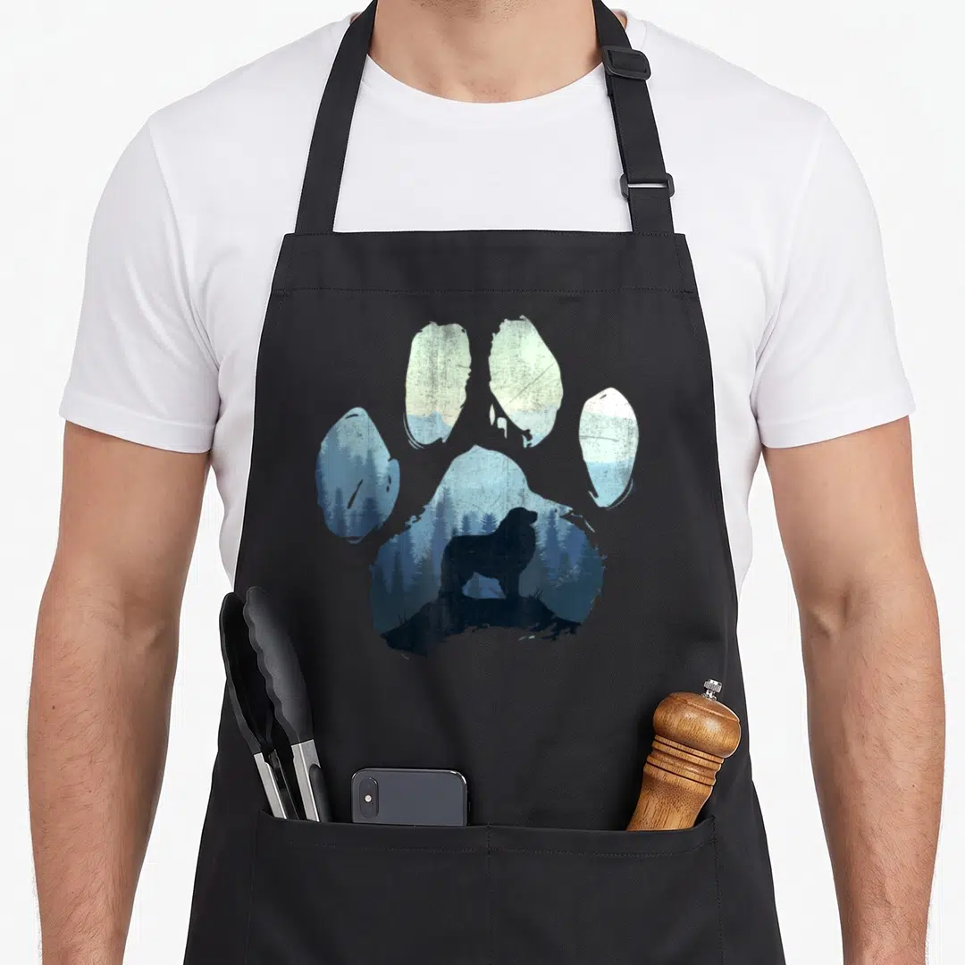 Pyrenee paw dog apron - Paw print silhouette containing a bear standing on a rock in a blue forest landscape — shown on a black cotton canvas apron against a white background