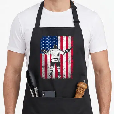 Ice hockey american flag apron - Distressed white hockey player silhouette holding stick across shoulders against red — shown on a black cotton canvas apron against a white background