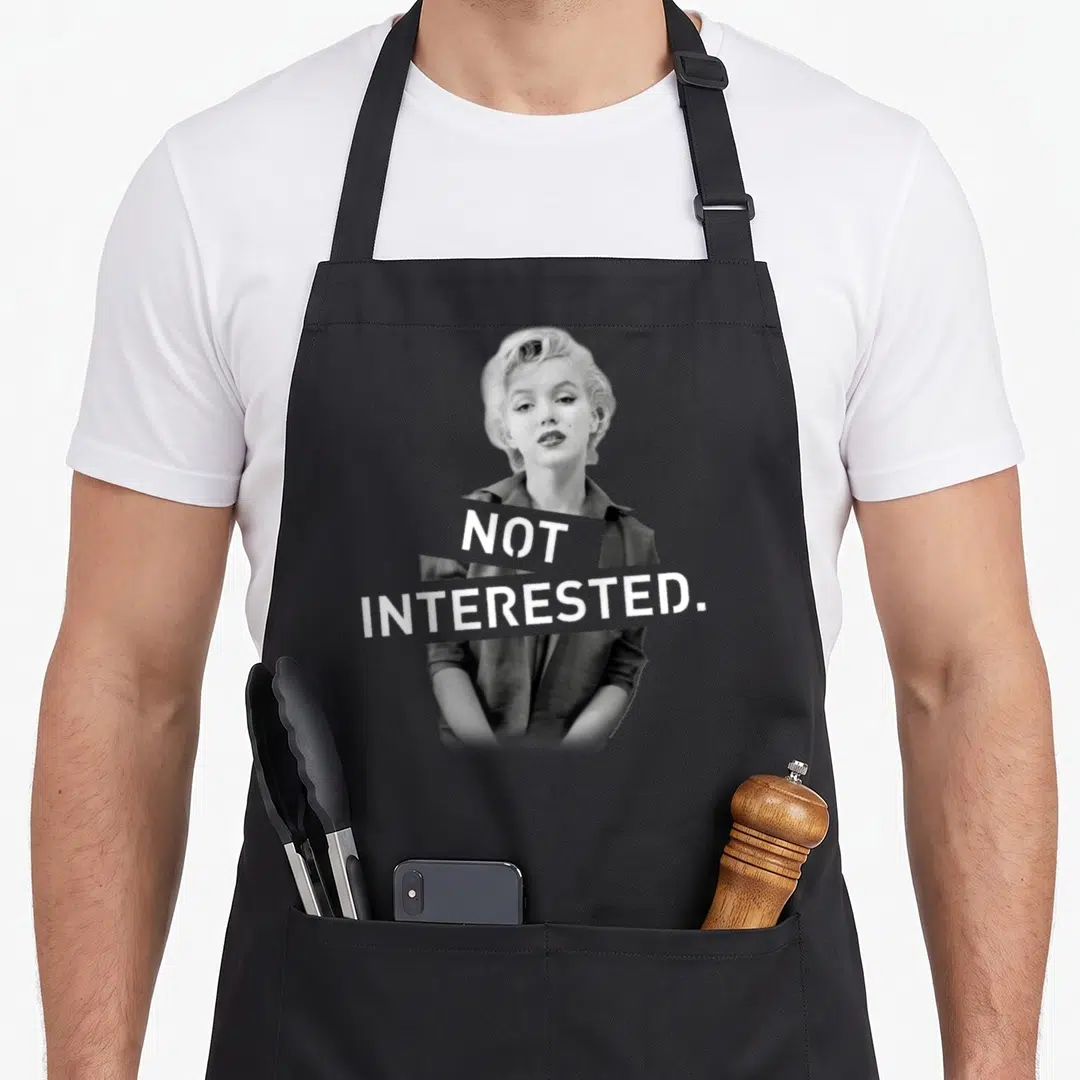 Artwork marilyn monroe apron - Grayscale Marilyn Monroe photo with white NOT INTERESTED text on grey blocks — shown on a black cotton canvas apron against a white background