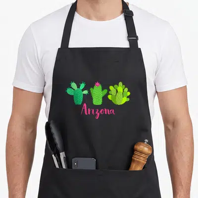 Flower cactus apron - Three green cacti and pink Arizona script on black fabric — shown on a black cotton canvas apron against a white background