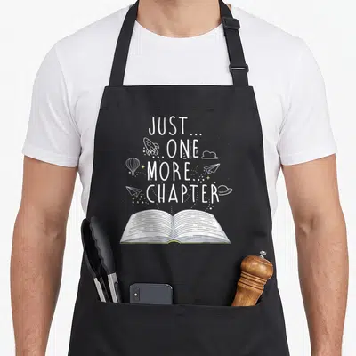 Chapter reading book apron - Just One More Chapter text with floating rocket, planes and open book line art — shown on a black cotton canvas apron against a white background