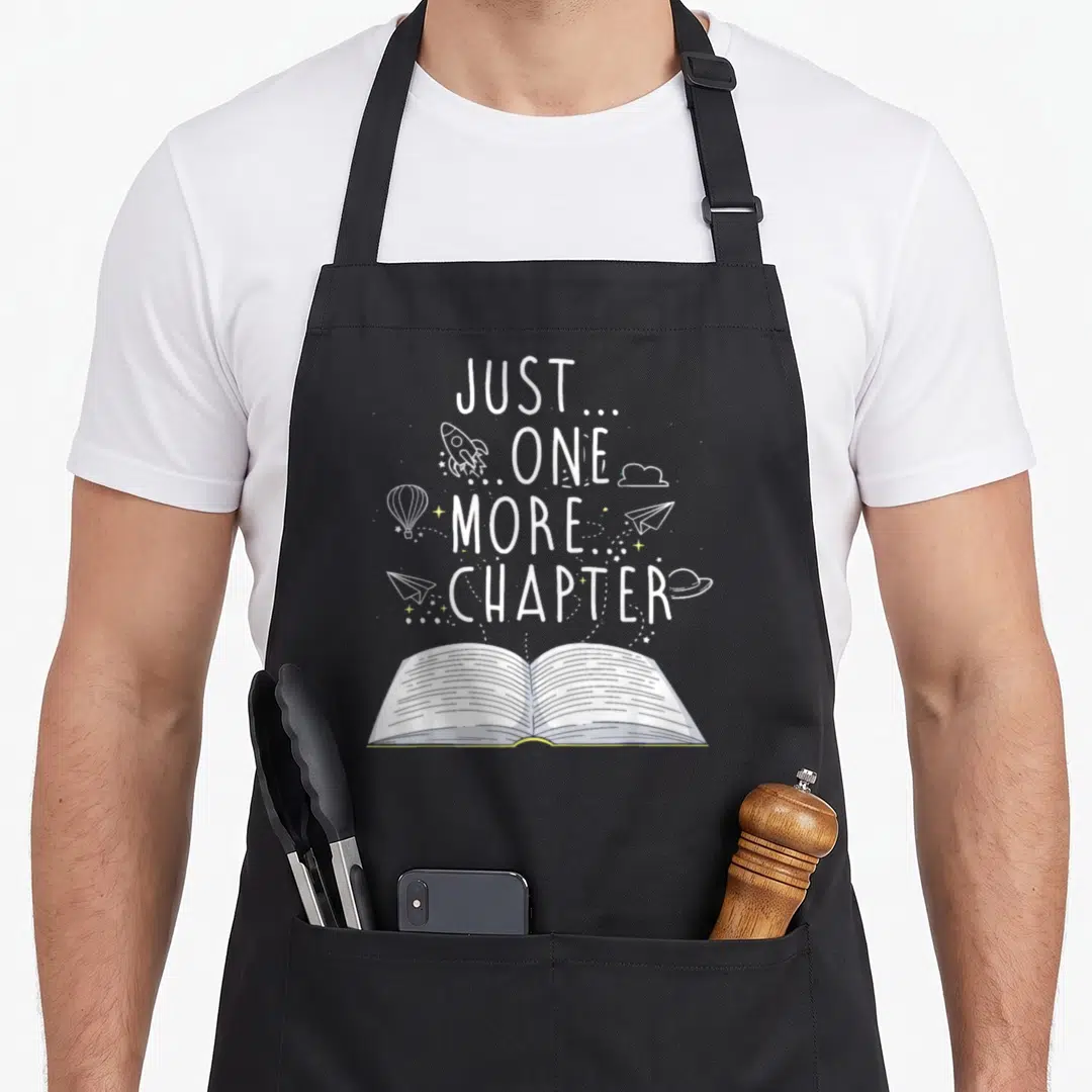 Chapter reading book apron - Just One More Chapter text with floating rocket, planes and open book line art — shown on a black cotton canvas apron against a white background