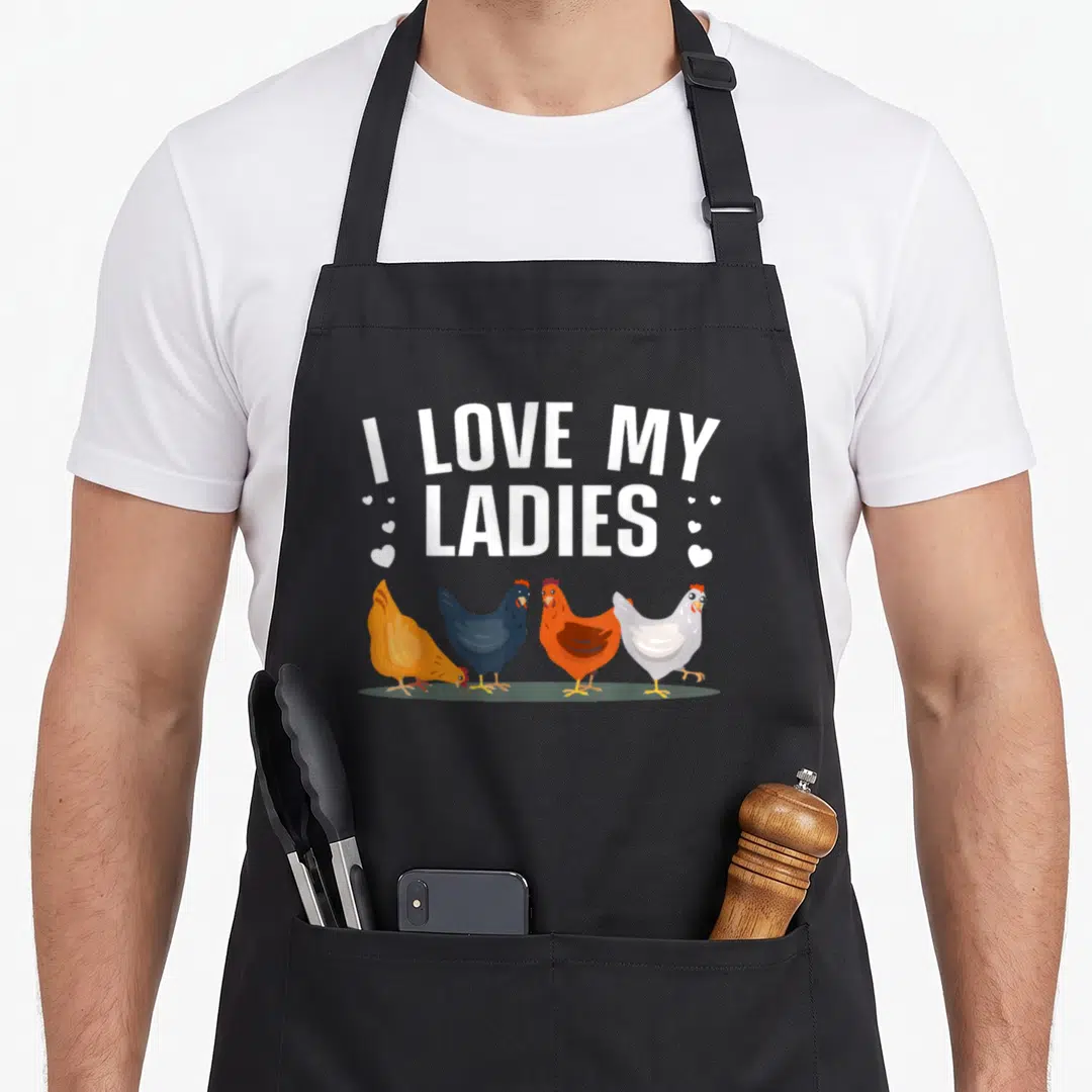 Farmer chicken with apron - I Love My Ladies text above four white yellow blue grey orange brown cartoon hens — shown on a black cotton canvas apron against a white background