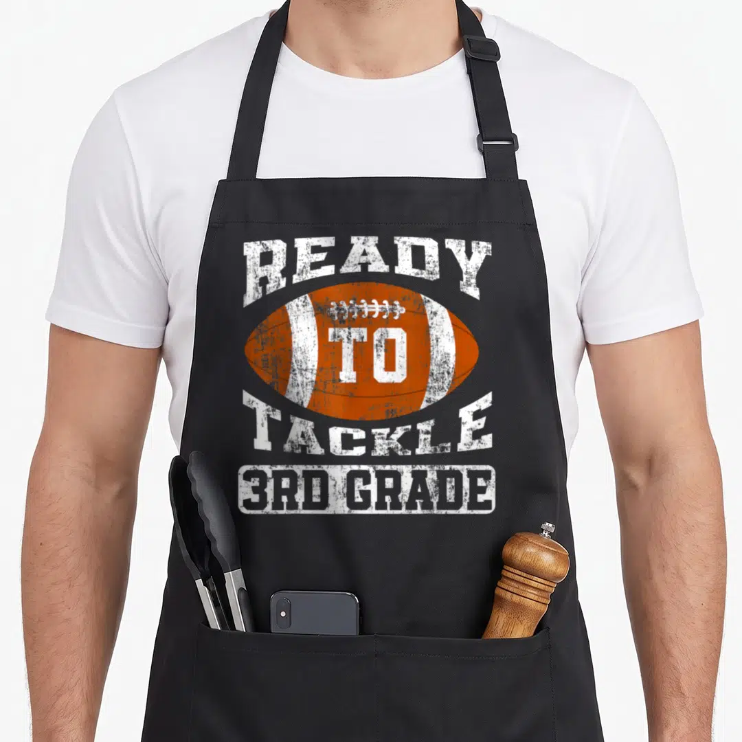 Ready football apron - Orange football with arched Ready To Tackle 3rd Grade text in distressed white font — shown on a black cotton canvas apron against a white background