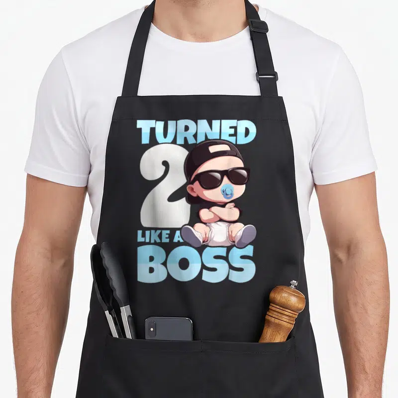 2nd bbq boss apron - Cartoon baby with sunglasses and cap over white number 2 with blue text — shown on a black cotton canvas apron against a white background