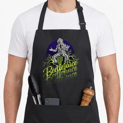Funny three novelty apron - Beetlejuice sitting on tombstone with purple circle and three lines of yellow text — shown on a black cotton canvas apron against a white background