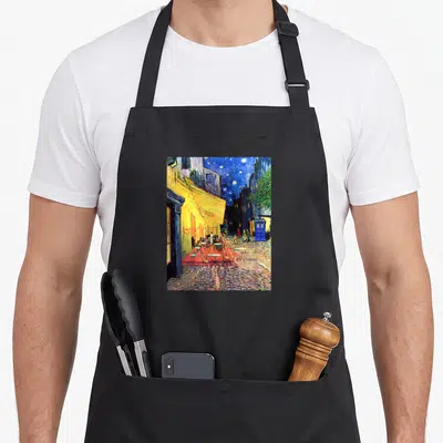 Cafe doctor apron - Street scene with yellow building, red tables, and blue phone booth under stars — shown on a black cotton canvas apron against a white background