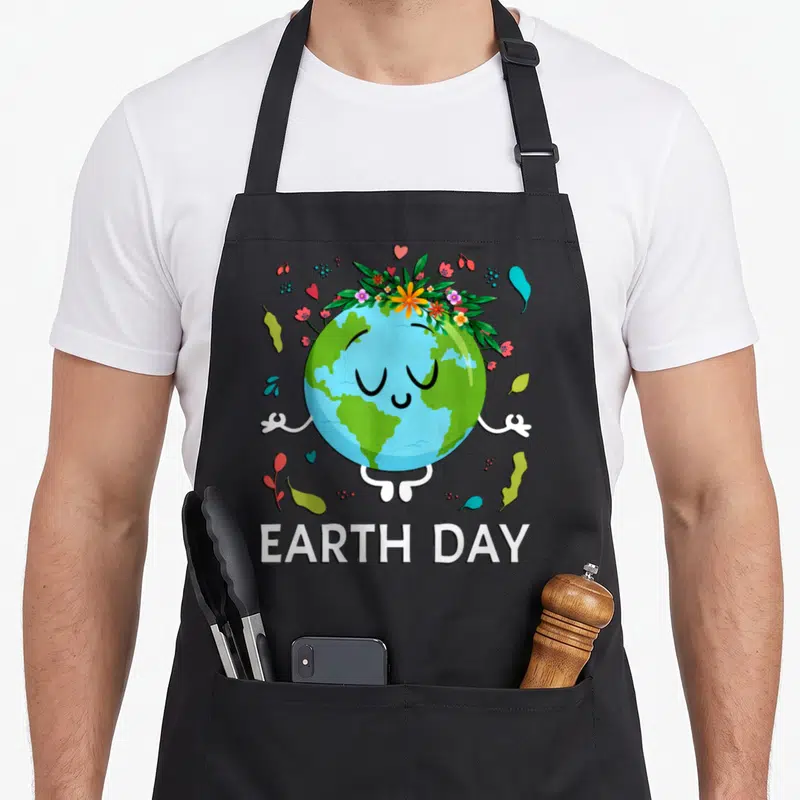 Peaceful floral apron - Cartoon Earth with flower crown doing yoga pose above EARTH DAY text — shown on a black cotton canvas apron against a white background