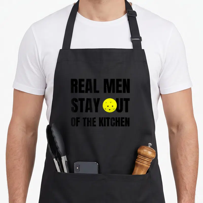 Funny stay pickleball apron - Black text REAL MEN STAY OUT OF THE KITCHEN with yellow perforated ball replacing O in OUT — shown on a black cotton canvas apron against a white background