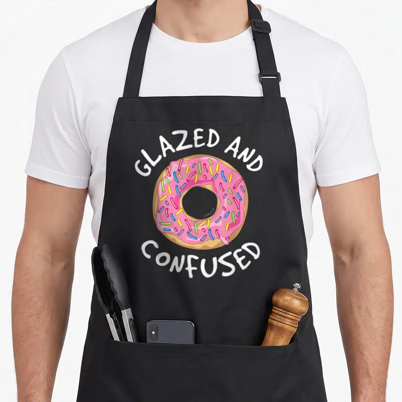 Glazed donut apron - Pink frosted donut with sprinkles and white text reading GLAZED AND CONFUSED — shown on a black cotton canvas apron against a white background