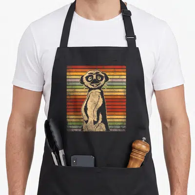 Meerkat apron for mom - Retro grey meerkat standing in front of red orange yellow green purple and grey horizontal — shown on a black cotton canvas apron against a white background