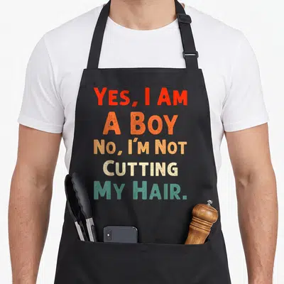 Cute hair cutting apron - Stacked red orange beige and teal text reading Yes I Am A Boy No I'm Not Cutting My Hair — shown on a black cotton canvas apron against a white background