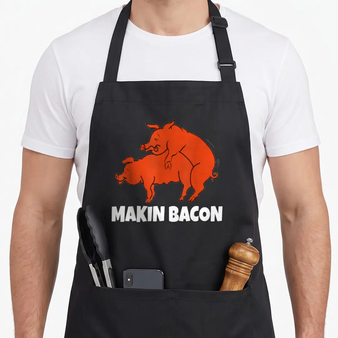 Smoking bacon apron - Two orange cartoon pigs mating above white MAKIN BACON text — shown on a black cotton canvas apron against a white background