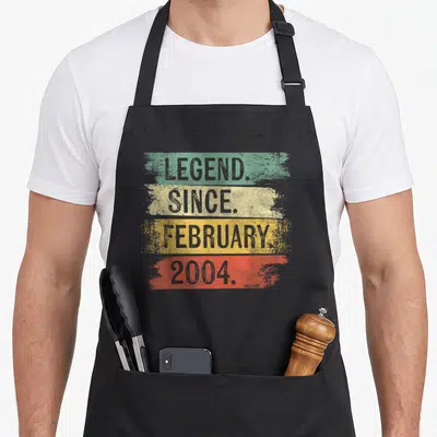 Retro legend birthday apron - Stacked text LEGEND SINCE FEBRUARY 2004 over green yellow orange and red stripes — shown on a black cotton canvas apron against a white background