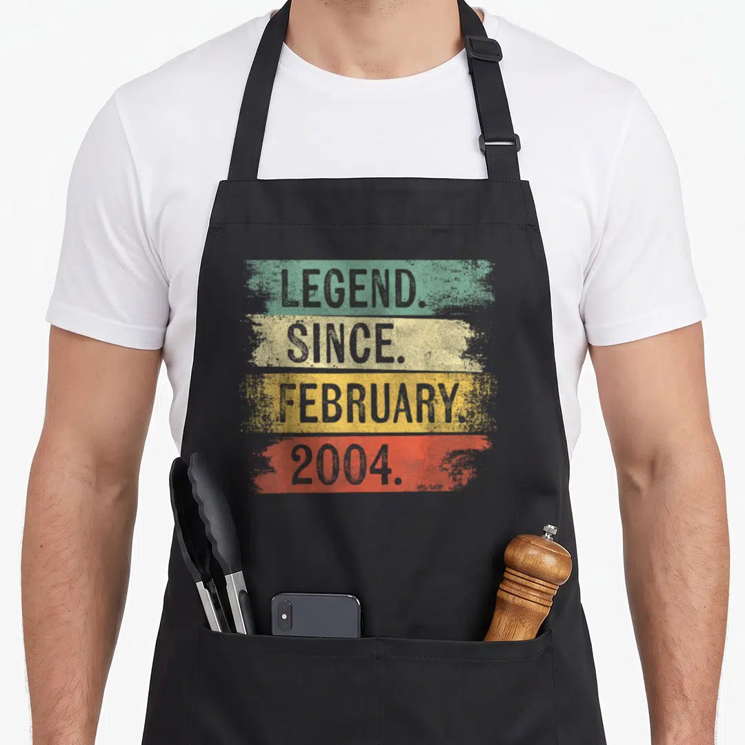 Retro legend birthday apron - Stacked text LEGEND SINCE FEBRUARY 2004 over green yellow orange and red stripes — shown on a black cotton canvas apron against a white background